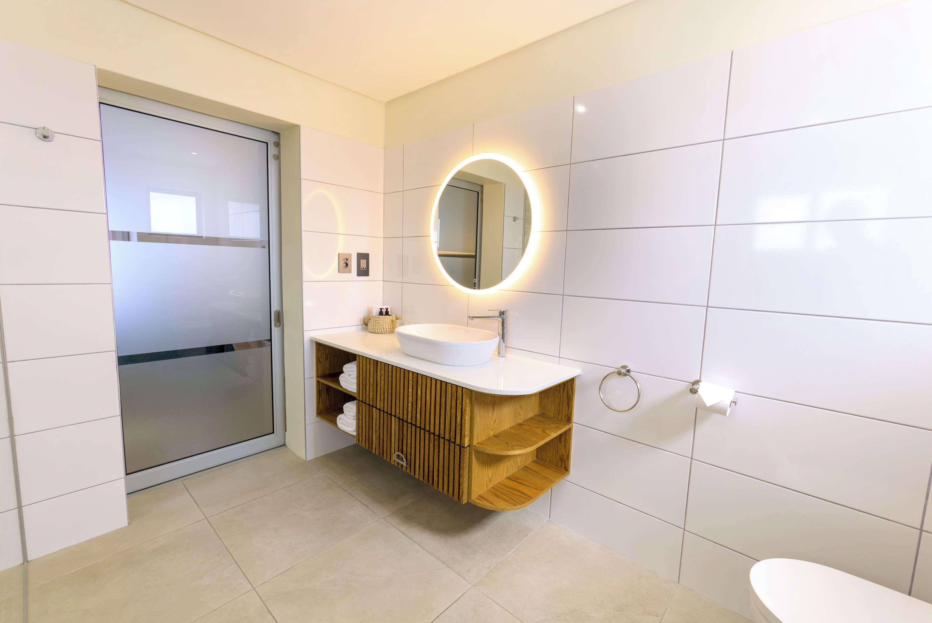 A modern bathroom with a glass door entrance, a white sink with a wooden vanity, and a round mirror with a light.