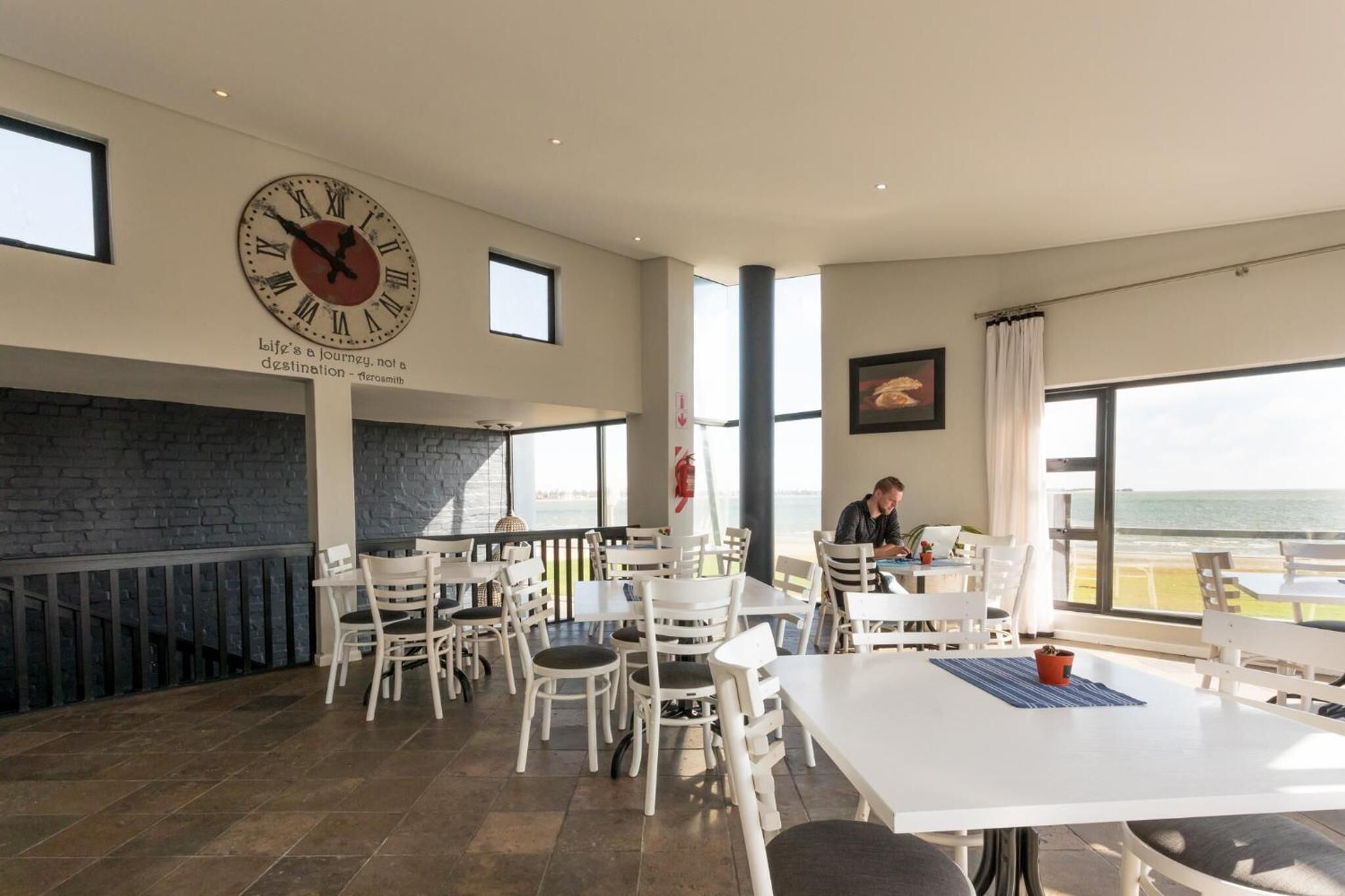 Un comedor amplio con mesas y sillas blancas, un reloj de pared grande y vista al mar.