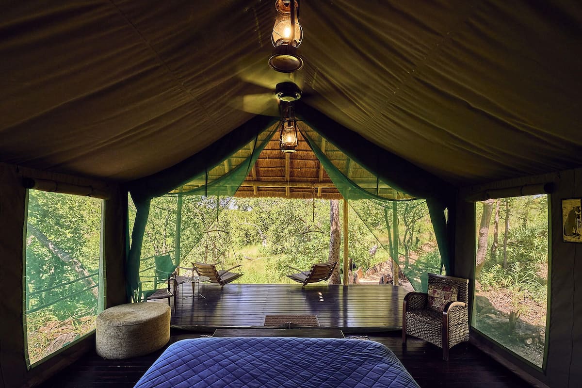 A tented room with a bed, two chairs, a hanging light fixture, and large windows offering a forest view.