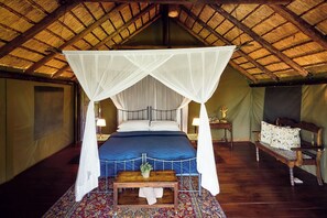 A four-poster bed with a canopy, a wooden nightstand with a lamp, a small table with a plant, and a patterned rug. - Parsons Hilltop Safari Camp (Hoedspruit)