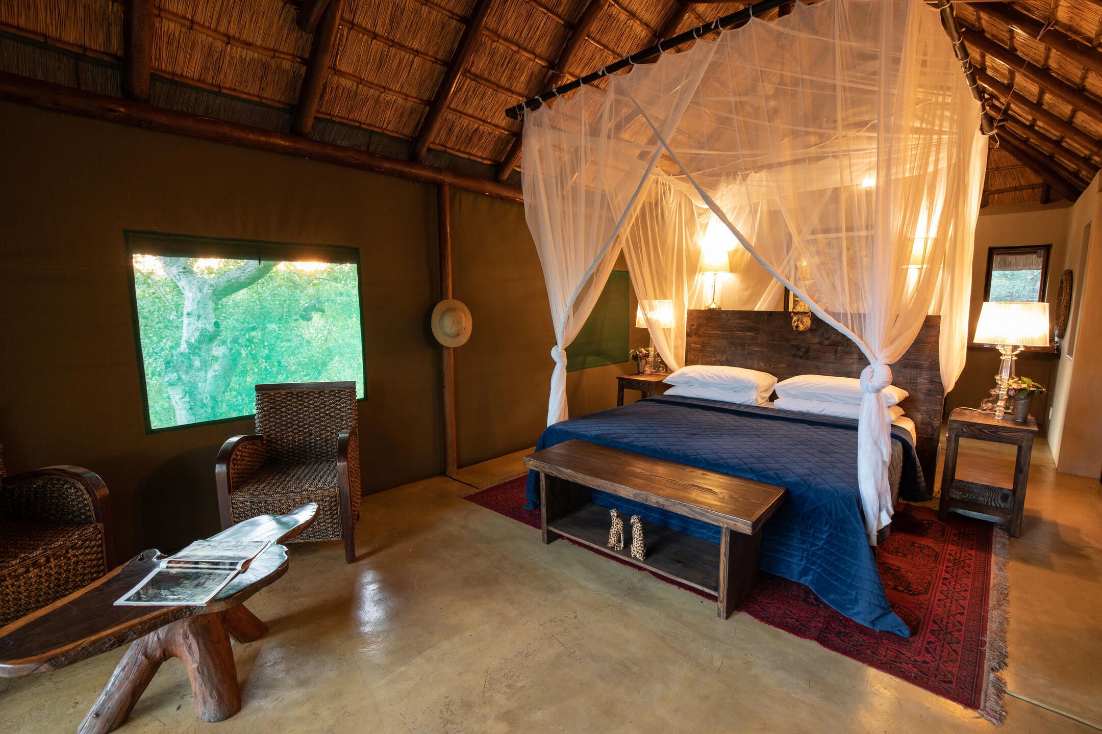 A room with a bed, a chair, a small table, a nightstand, and a window with a view of greenery.