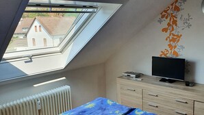 Ein Schlafzimmer mit Oberlicht, einem Bett mit blauer Decke, einer hölzernen Kommode und einem Fernseher.