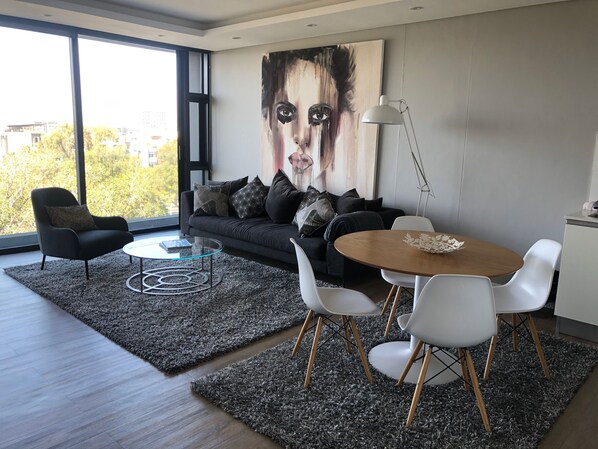 A modern living room with a dark sofa, a glass coffee table, white chairs, and a large abstract painting on the wall.