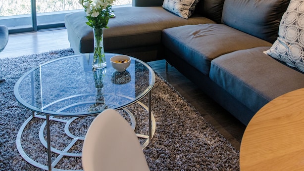 Uma sala de estar moderna com uma mesa de centro de vidro, um sofá com almofadas estampadas e vista para a cidade através de janelas amplas.