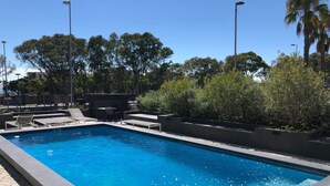 Uma piscina retangular com água azul cristalina, rodeada por espreguiçadeiras e uma pequena mesa.