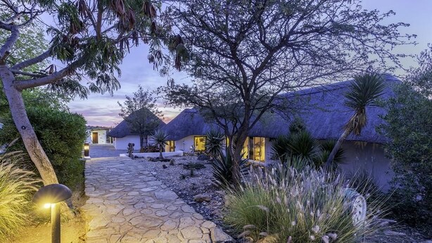 A pathway leading to thatched-roof cottages with outdoor lighting, surrounded by native vegetation. - Trans Kalahari Inn (Windhoek)