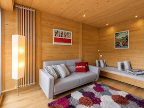 A modern living room with a grey sofa, red and grey cushions, a colorful rug, wooden walls, and two wall-mounted pictures.