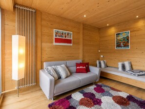 Una sala de estar moderna con un sofá gris, cojines rojos y grises, un tapete colorido, paredes de madera y dos cuadros colgados.