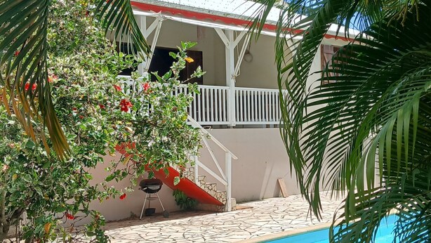 A pool area with a tiled deck, a covered patio, and a view of a building with a white railing and red accents. - Creole villa with pool in tropical garden   (Sainte Rose)