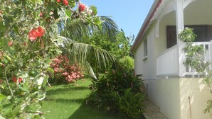 A garden with various plants and flowers, a white building with a balcony, and a stone pathway. - Creole villa with pool in tropical garden   (Sainte Rose)