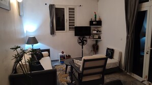 Un espacio habitable compacto con un sofá, una mesita, un ventilador y un televisor sobre una estantería.
