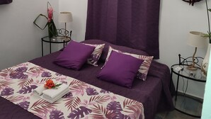 Un dormitorio con cama, ropa de cama morada, dos mesitas de noche con lámparas y dos sombreros en la pared.