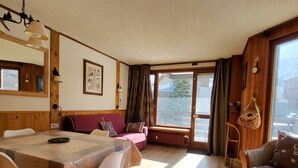 A cozy room with a wooden dining table, a sofa, and a wicker chair. There is a window with curtains and a view of the outside. - Tignes: 2-room apartment 41m², 500m from slopes, southwest balcony, wifi, 4-5 pers. (Tignes)