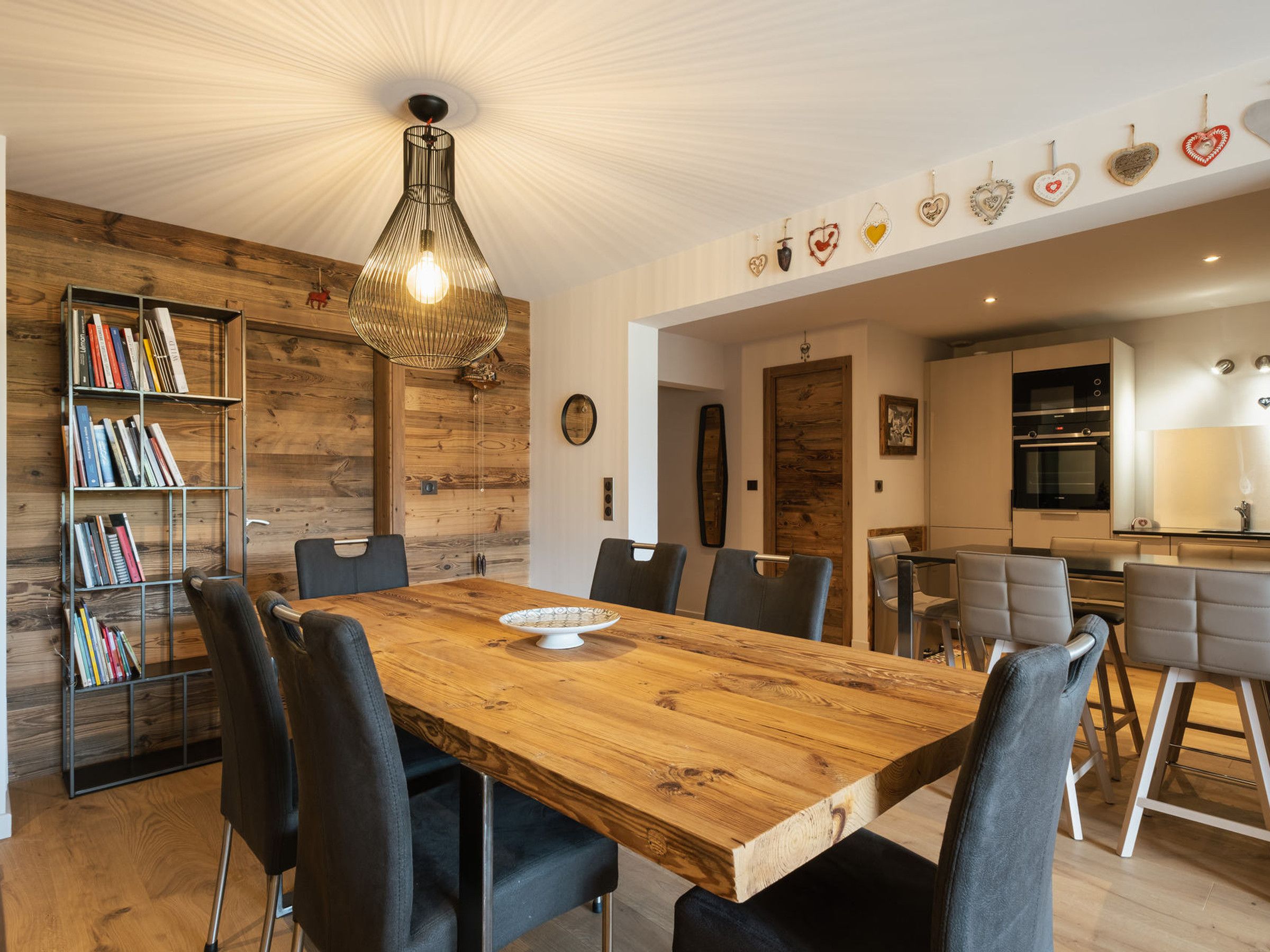 Uma sala de jantar com mesa de madeira, cadeiras, estante e luminária pendente.