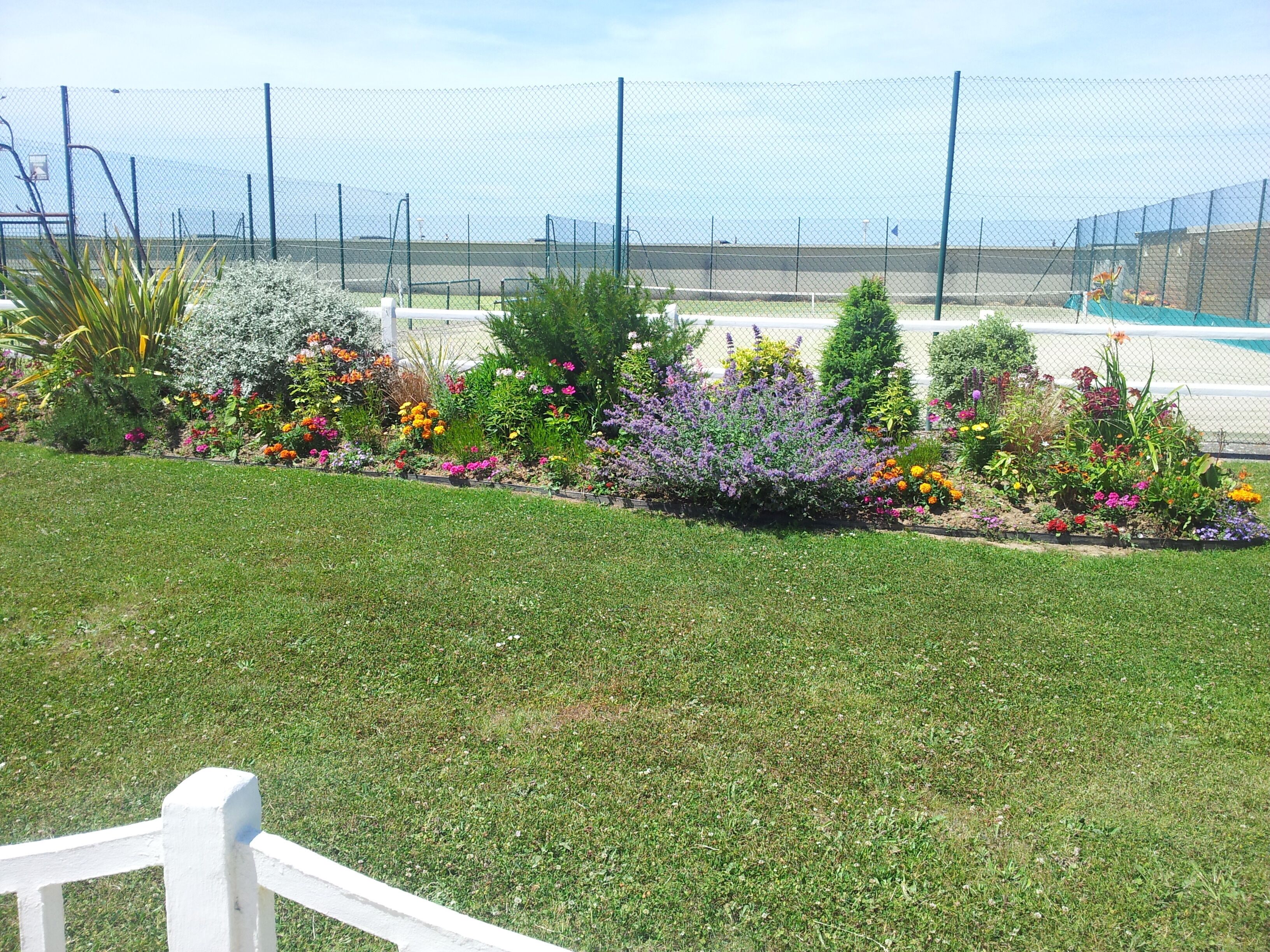 A well-maintained garden with a variety of colorful flowers and shrubs, a white fence, and a sports field in the background.
