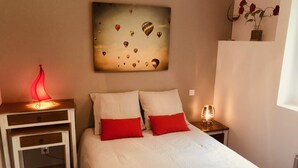 Ein Schlafzimmer mit einem Bett, zwei Nachttischen, einem Schminktischchen, einem Wandbild mit Heißluftballons und einer Blumenvase auf der Kommode.