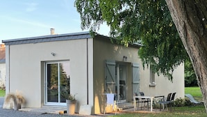 Ein modernes Gebäude mit Glastür und -fenstern, eine kleine Terrasse mit Tisch und Stühlen sowie ein Baum im Vordergrund.