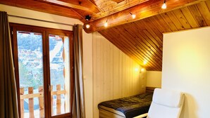 Una habitación acogedora con cama, una silla, pisos de madera, techo de madera y una ventana con cortinas.