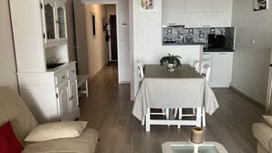 Un salon moderne comprenant un canapé, une table à manger avec des chaises, un coin cuisine avec des meubles blancs et un couloir menant à une autre pièce.