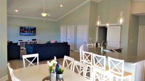 A modern living room with a dark blue sofa, a dining table with white chairs, and a kitchen area with a granite countertop and pendant lights. - GREAT OCEAN VIEWS -- 5 Bedroom, 5.5 Bath, 2 Living Rooms (Ocean Isle Beach)