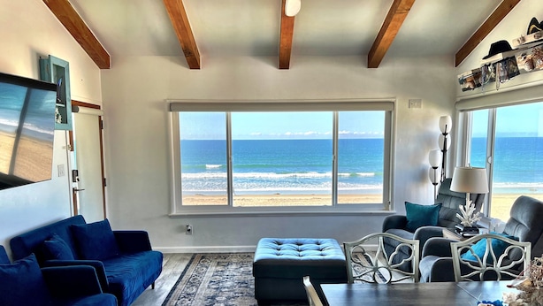 Ein Zimmer mit großem Fenster zum Strand, ein blaues Sofa, ein Esstisch mit Stühlen und ein Deckenventilator.