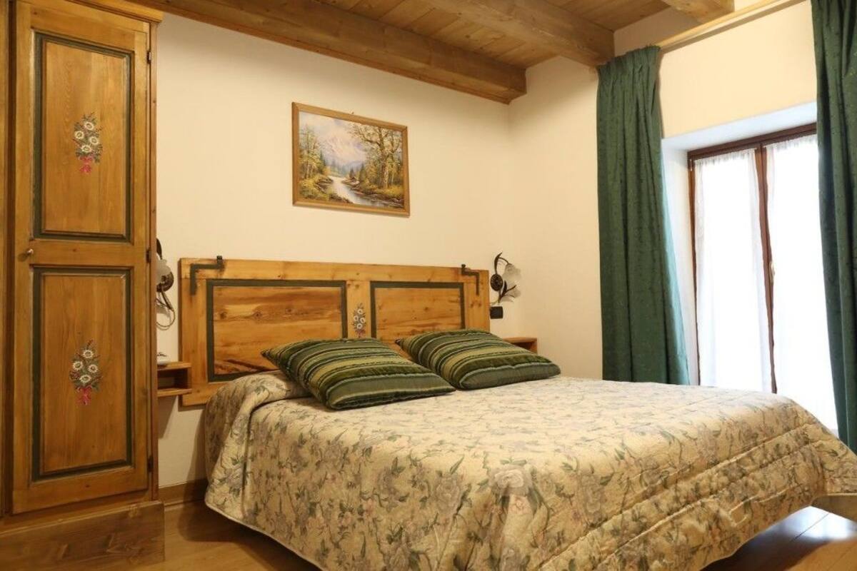Un dormitorio con cabecera de madera, dos almohadas verdes a rayas, colcha estampada con motivos florales, un cuadro con paisaje de bosque y una ventana con cortinas verdes.