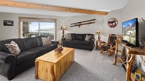 Una sala de estar moderna con dos sofás oscuros, una mesa de centro de madera y un televisor. Hay sillas de madera y una lámpara sobre una mesita auxiliar. Un soporte para esquíes está montado en la pared, y un ventanal ofrece vistas al exterior.