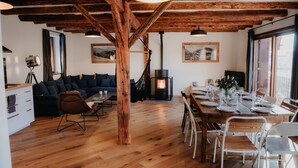 Een ruime woonkamer met houten plafond, een zithoek, een eettafel voor zes personen en een open haard.