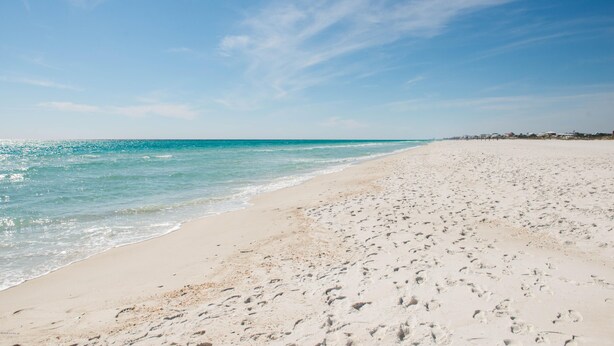 A wide sandy beach with footprints leading to the water, a clear blue sky, and distant buildings.
