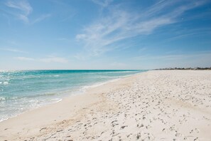 Une vaste plage de sable où l’on peut voir des empreintes de pas menant à l’eau, un ciel dégagé et une côte lointaine parsemée de bâtiments.