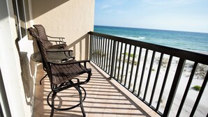 A balcony with two wicker chairs and a table, overlooking a beach with waves and sand.