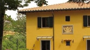 A yellow building with a tiled roof, a door, and windows. There is outdoor furniture and a potted plant. - Barco Mediceo (Carmignano)