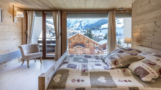 Ein Schlafzimmer einer Holzhütte mit einem Bett, einem Sessel und Blick auf ein Bergdorf.
