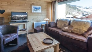 Ein gemütliches Blockhäuschen im Inneren, mit Holzbalkendecke, einem an einem hölzernen Standfuß befestigten Fernseher, einer Couch mit kuscheligen Fellkissen und Blick auf eine verschneite Berglandschaft durch ein großes Fenster.