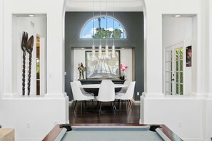 A dining area with a pool table, white chairs, and a large window with a view of palm trees.