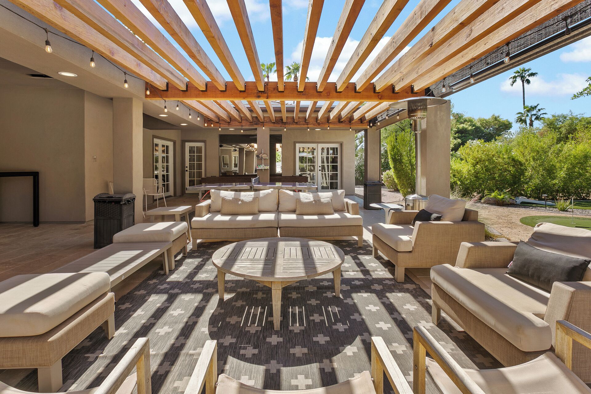 Een terras buiten met een houten pergola, banken, een ronde tafel en een kamerkleed met een patroon.