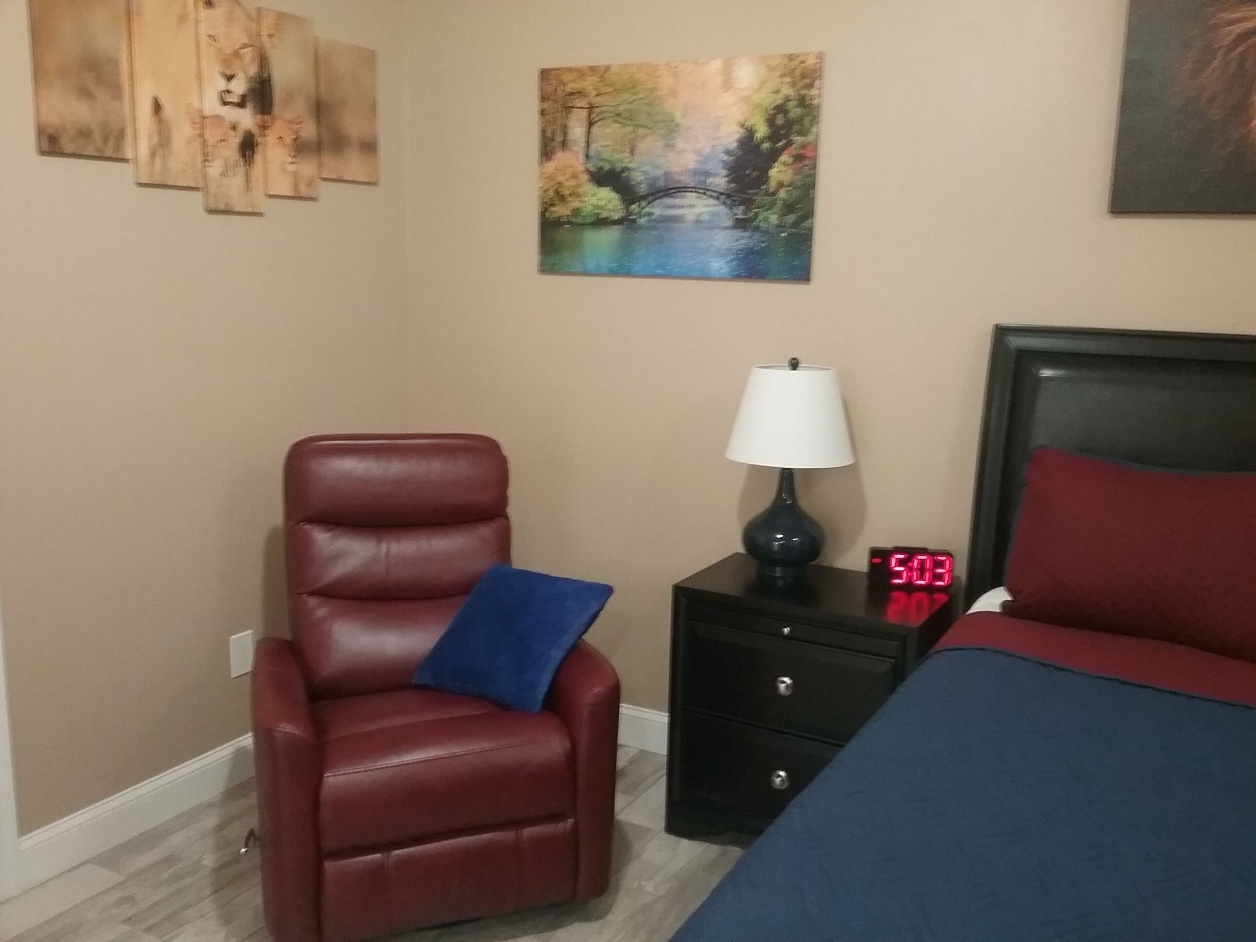 Una habitación de hotel con un sillón de cuero rojo, una mesita de noche con lámpara, un reloj digital y una cama con almohada roja.