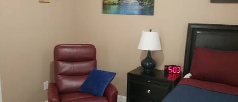 Una habitación de hotel con un sillón de cuero rojo, una mesita de noche con lámpara, un reloj digital y una cama con almohada roja.
