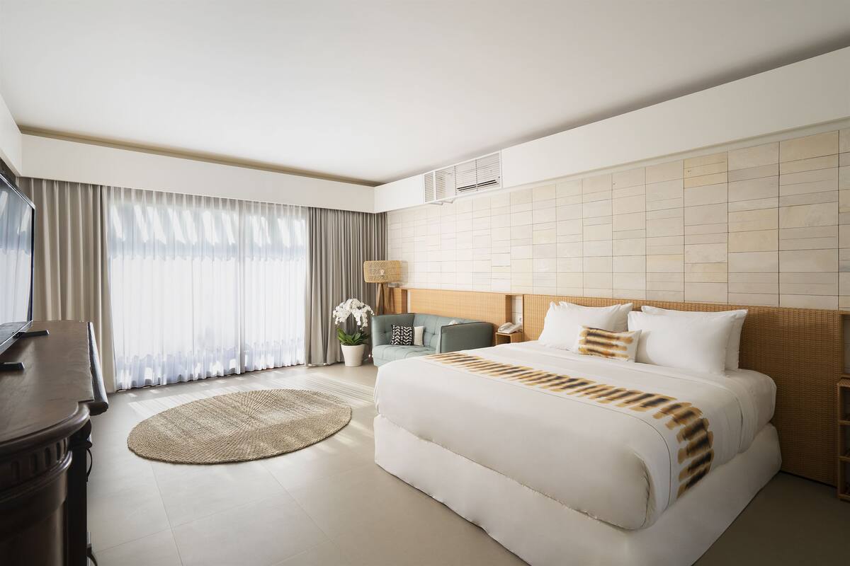 A hotel room with a large bed, a desk, a chair, a potted plant, and a view of the city through the window.