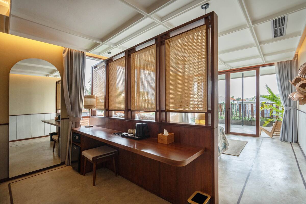 A hotel room with a wooden desk, a small seating area, a large window with a view, and a balcony.