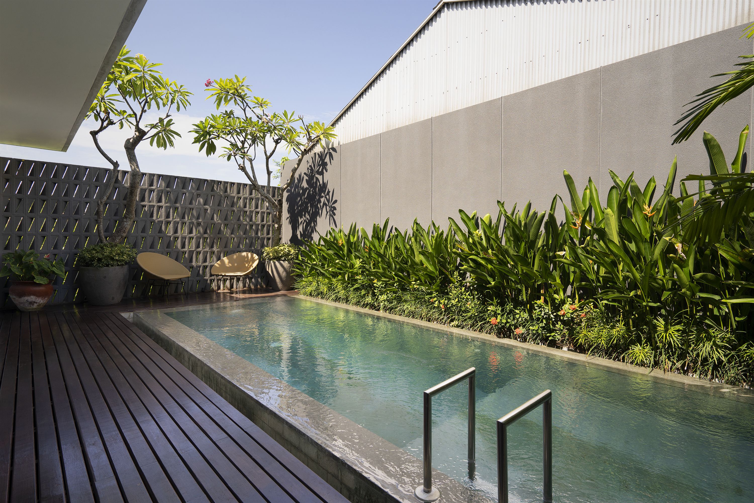 A swimming pool with a wooden deck, surrounded by lush greenery and a modern building.