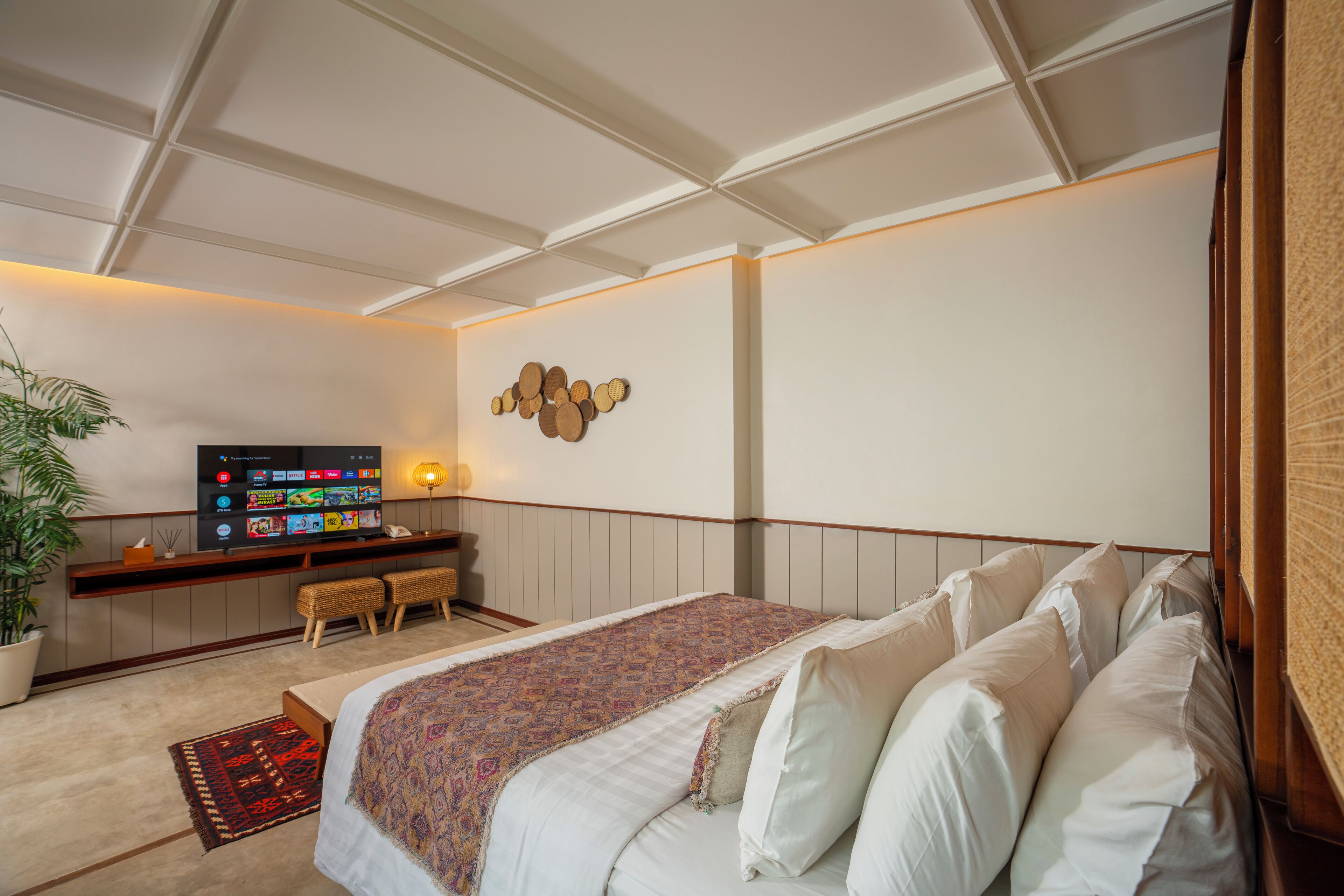 A bedroom with a large bed, a TV on a stand, a potted plant, and a wall-mounted light fixture.