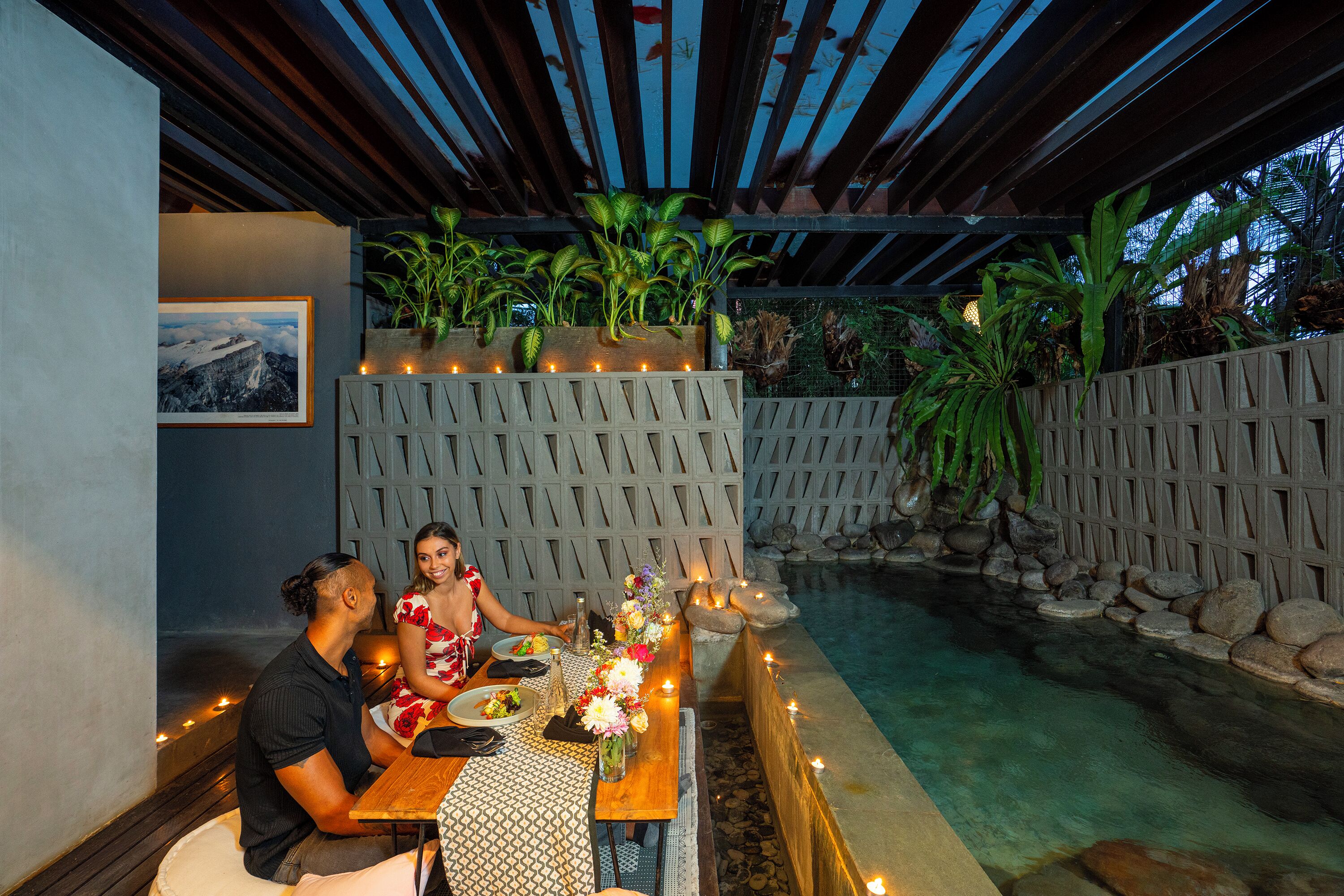 A dining area with a pool, wooden table, candles, and plants.