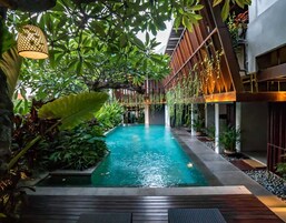 Un área de piscina con cubierta de madera, rodeada de abundante vegetación y una estructura de edificio moderna.