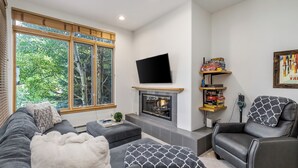 Ein modernes Wohnzimmer mit Kamin, grauen Sofas, einem schwarzen Ledersessel und einem großen Fenster mit Blick auf Bäume.