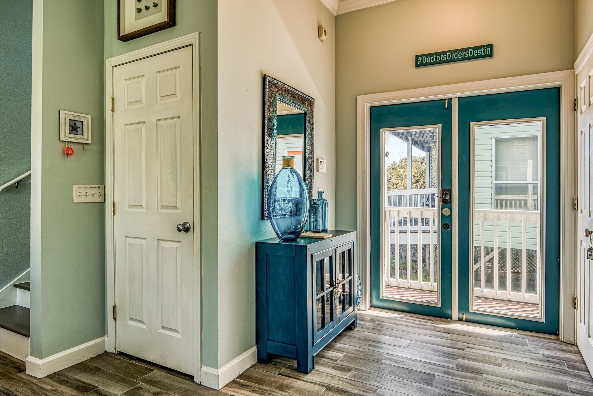 Un couloir avec une porte blanche, une console bleue ornée d’un miroir et une porte turquoise donnant sur un balcon.