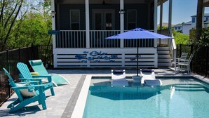 A swimming pool with lounge chairs and an umbrella, surrounded by a fence and a building with a porch.