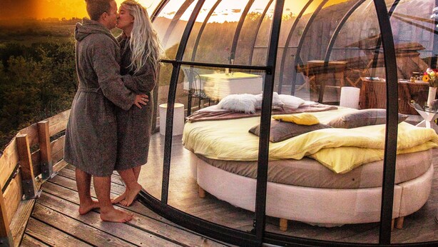 Un couple s’embrasse à l’intérieur d’une structure en forme de dôme, où l’on aperçoit un lit et une table.