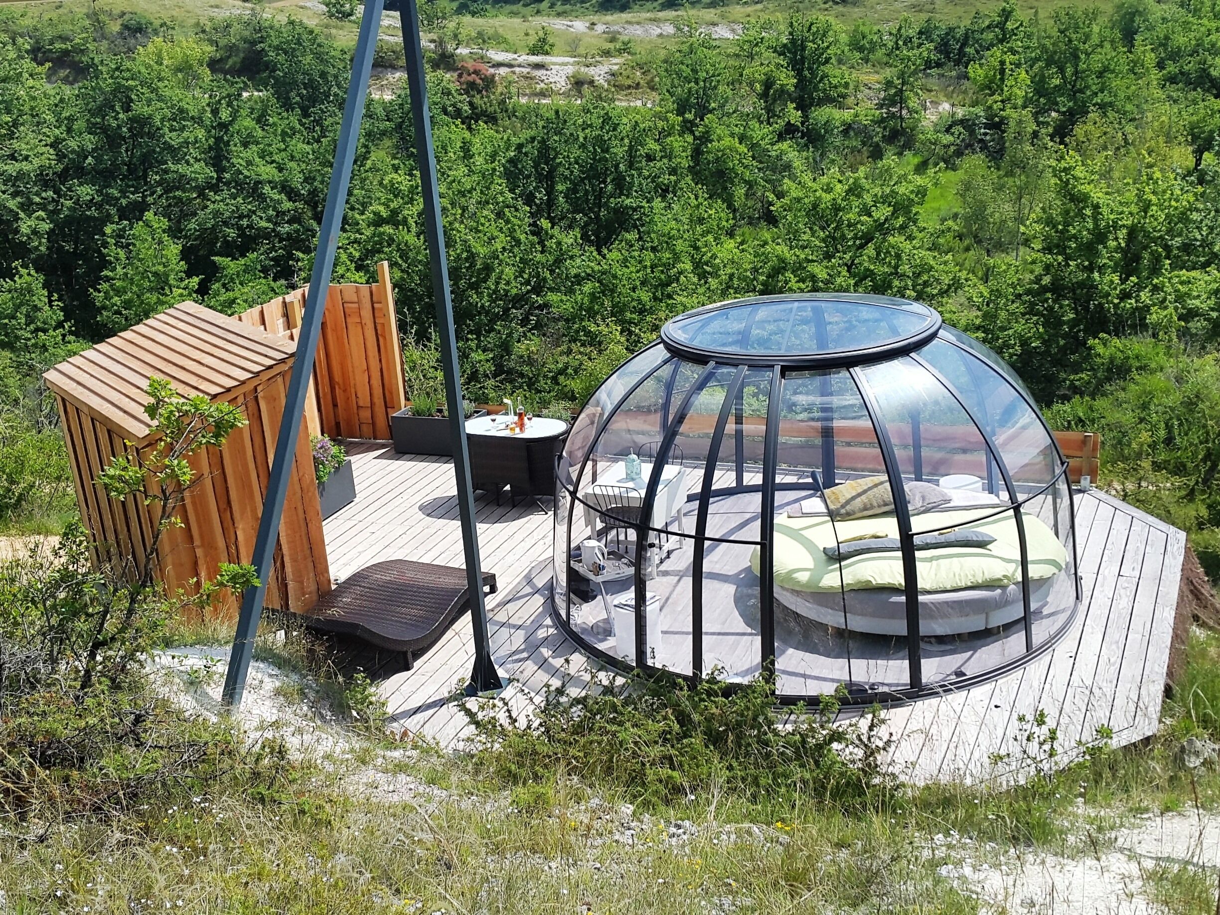 Une structure en forme de dôme transparent, avec une table ronde et des chaises à l’intérieur, entourée d’une terrasse en bois et d’un chalet en bois.