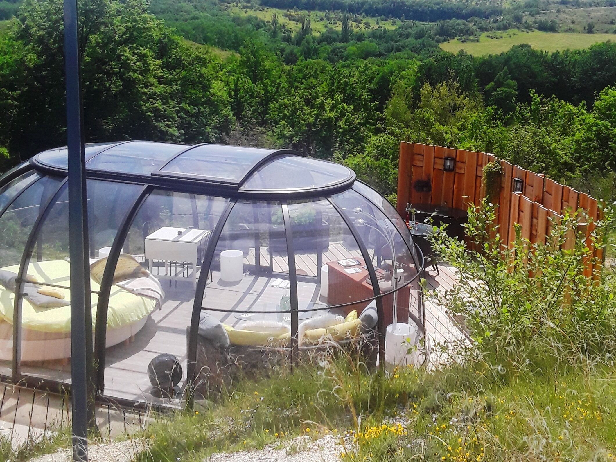 A transparent dome structure with a table and chairs inside, surrounded by a wooden deck and a wooden fence.